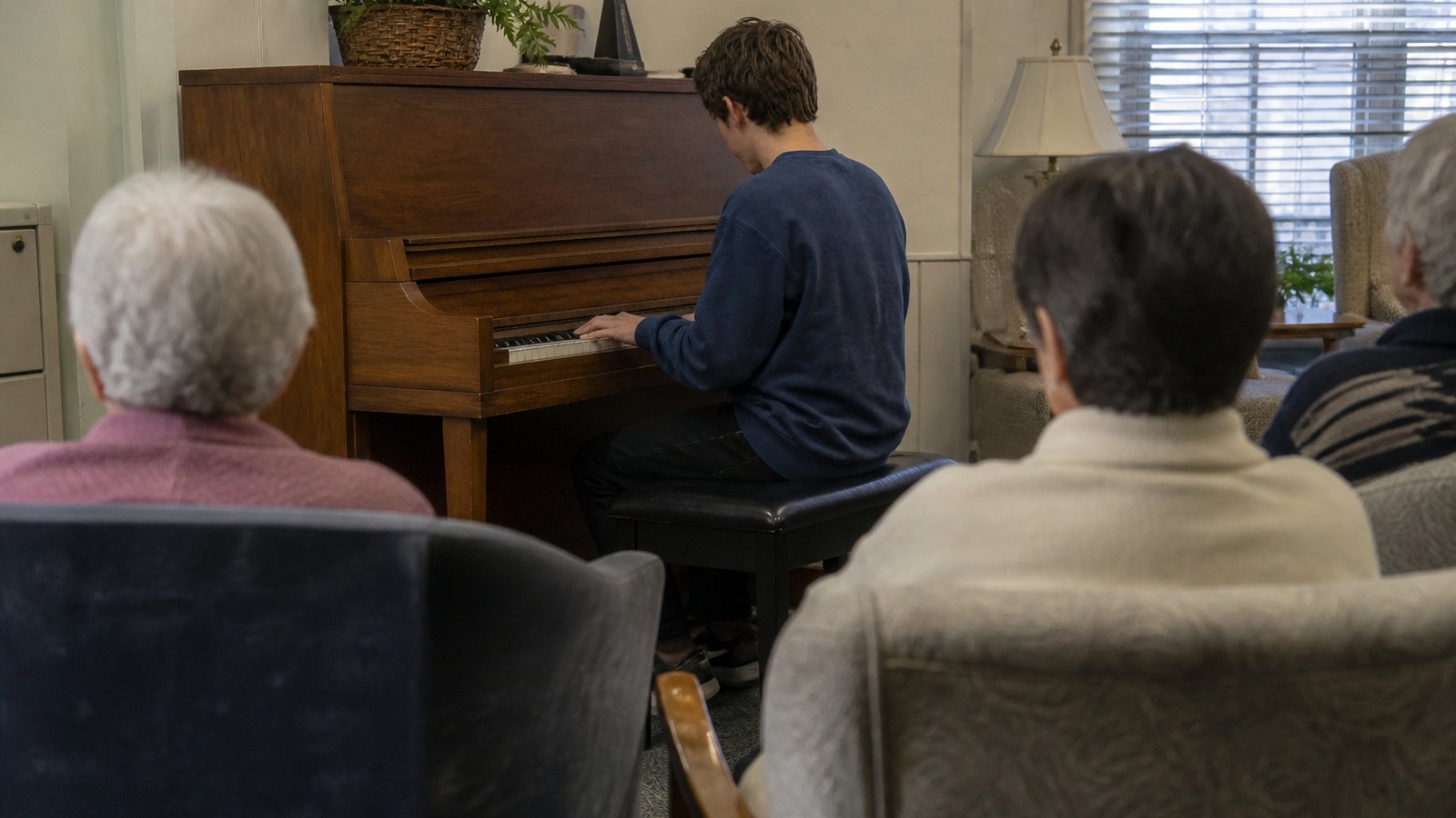 A student playing an upright piano for residents in a day room.